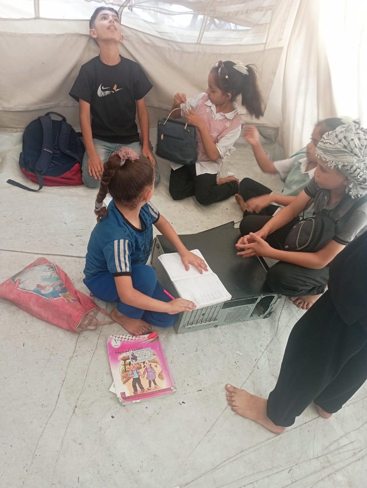 Children sitting on the floor of an old tent using a broken computer case as a desk